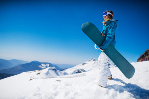 young woman stands on top of mountain holding a snowboard